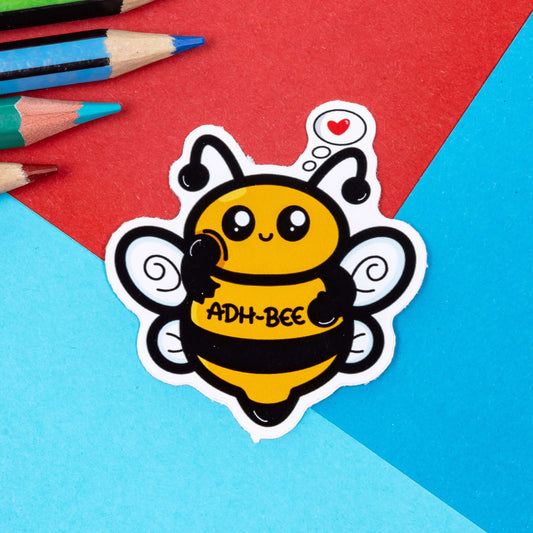 The ADH Bee Sticker - ADHD on a red and blue background with colouring pencils. The smiling bee sticker has a hand poking its cheek and a red heart thought bubble, across its middle in black reads 'ADH-Bee'. The hand drawn design is raising awareness for adhd and neurodiversity.