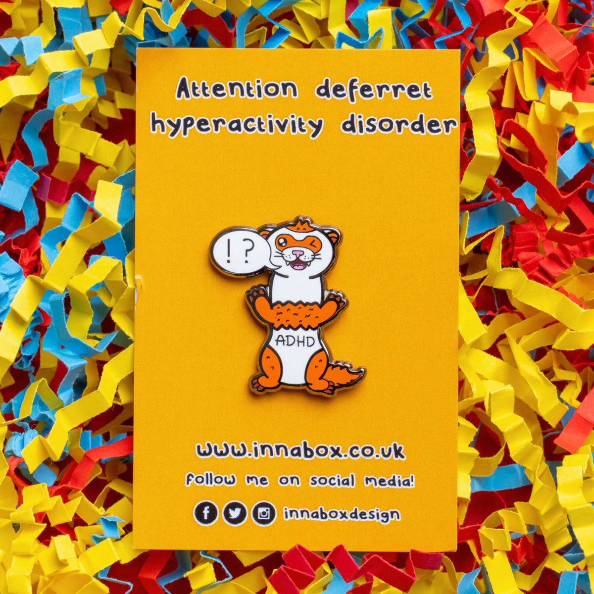 The Attention Deferret Hyperactivity Disorder Ferret Enamel Pin - ADHD on yellow backing card laid on a red, yellow and blue card confetti background. Red and white ferret pin smiling and winking with a speech bubble that has !? inside it and across its middle reads 'ADHD'. Hand drawn design is raising awareness for attention deficit hyperactivity disorder ADHD.