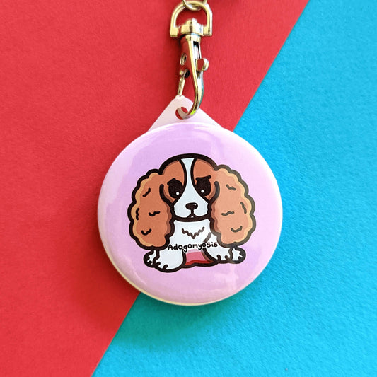 A sad spaniel dog with a red tummy and black text reading adogomyosis on a pink background plastic circular keychain hanging on a silver lobster clip. Adogomyosis Keyring - Adenomyosis is on a red and blue background. Raising awareness for adenomyosis.