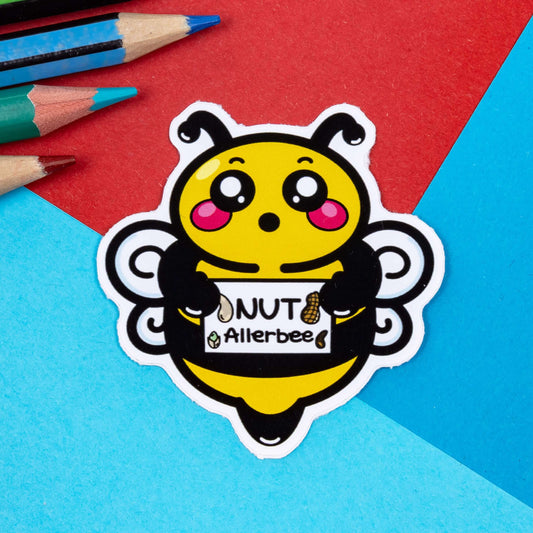 The Allerbee Bee Sticker - Nut Allergy on a red and blue background with colouring pencils. The bee sticker has a shocked expression with red cheeks holding up a sign reading 'nut allerbee' with different nuts. The hand drawn design is raising awareness for nut allergies.