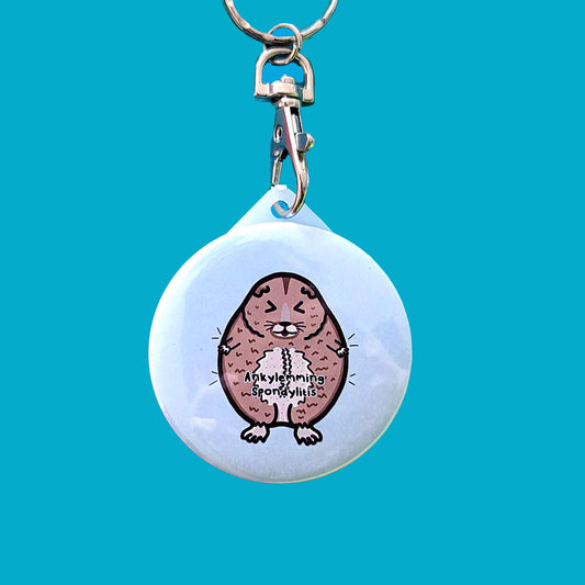 The Ankylemming - Ankylosing Spondylitis Keyring, a stressed lemming with its eyes scrunched shut on a white background circle keychain with the text ankylemming spondylitis being held up by a silver lobster clip on a blue background. Raising awareness for Ankylosing Spondylitis.