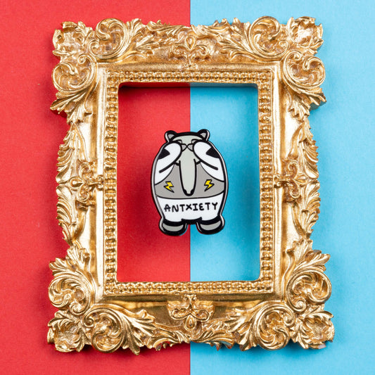 Antxiety Enamel Pin - Anxiety on blue and red card inside a gold ornate frame. The pin is a grey and white anteater with yellow lightning bolts and the word antxiety across its belly. The pin is designed to raise awareness for anxiety disorders and anxious emotions.