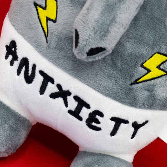 Close-up of the Antxiety Anteater Plush Toy by Innabox, showing the word “ANTXIETY” embroidered across its tummy in black text with lightning bolt details. A grey and white soft toy designed to represent anxiety with a cute and uplifting twist.