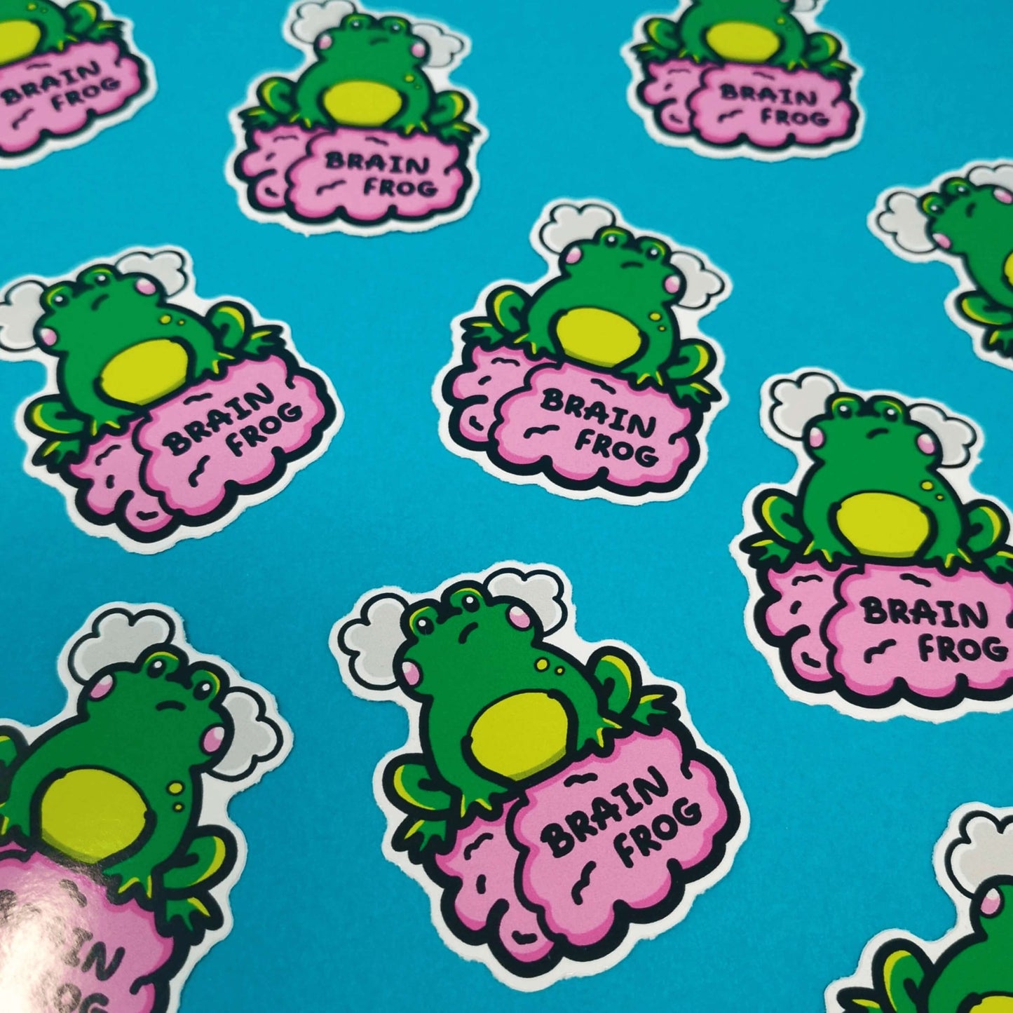 The Brain Frog Sticker - Brain Fog, multiple frog stickers on a blue background. The sticker is a confused frog with pink blush cheeks and two grey clouds above its head. Its sat on a pink brain with text that reads Brain Frog. The design was created to raise awareness for brain fog.
