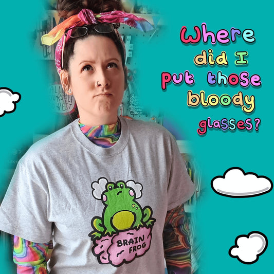 Nikky Box from innabox wearing a rainbow hair tie, a grey brain frog t-shirt - brain fog with a rainbow tie dye mesh long sleeve underneath. She is on a blue background with white clouds and rainbow text on her right reading 'Where did I put those bloody glasses?' with glasses on her head. The t-shirt print is a confused frog with pink blush cheeks and two grey clouds above its head. Its sat on a pink brain with text that reads Brain Frog. The design was created to raise awareness for brain fog.