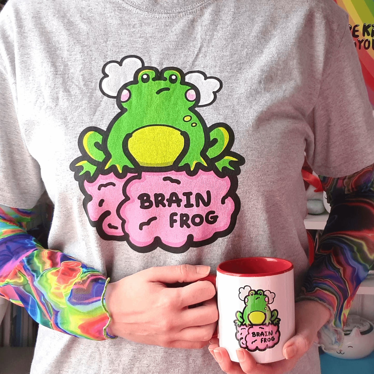 Close up of Nikky Box from innabox wearing a rainbow hair tie, a brain frog t-shirt - brain fog in grey with a rainbow tie dye mesh long sleeve underneath with a Brain Frog Mug - Brain Fog resting on her front. The print is a confused frog with pink blush cheeks and two grey clouds above its head. Its sat on a pink brain with text that reads Brain Frog. The design was created to raise awareness for brain fog.