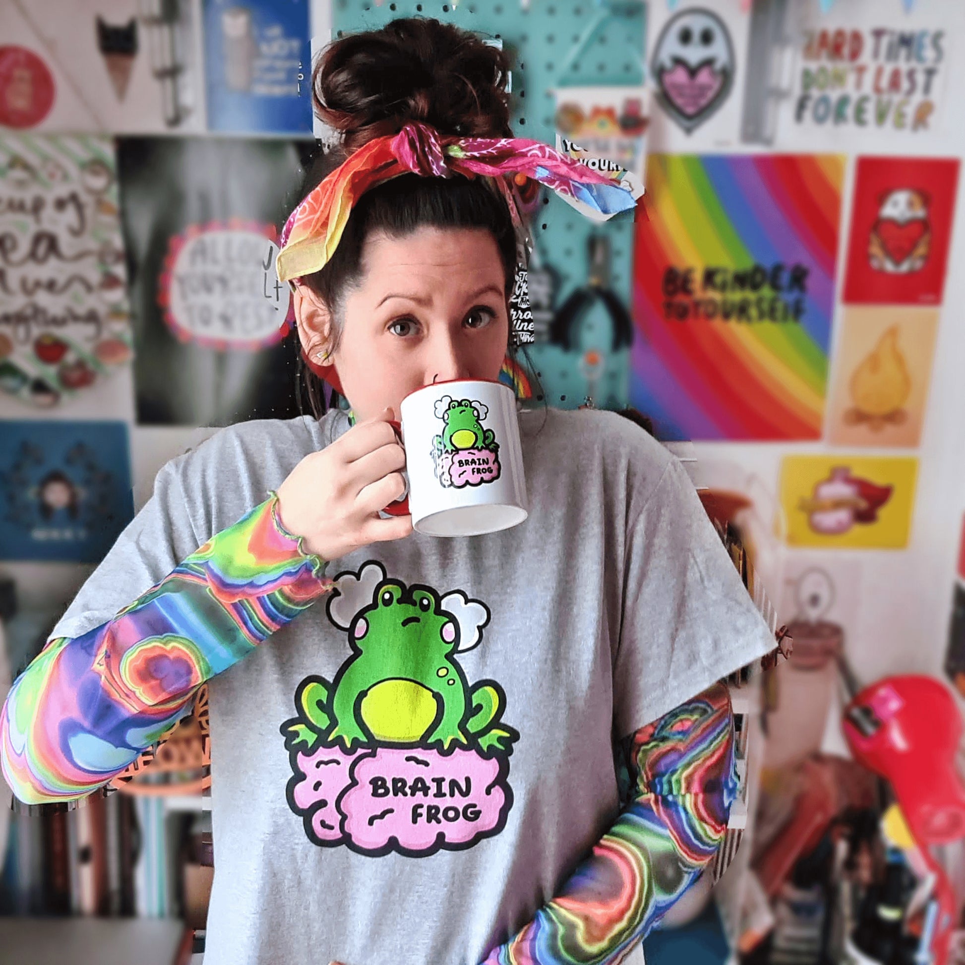Nikky Box from innabox wearing a rainbow hair tie, a grey brain frog t-shirt - brain fog with a rainbow tie dye mesh long sleeve underneath with a Brain Frog Mug - Brain Fog lifted to her mouth. The t-shirt print is a confused frog with pink blush cheeks and two grey clouds above its head. Its sat on a pink brain with text that reads Brain Frog. The design was created to raise awareness for brain fog.