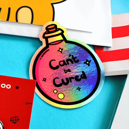 The Can't Be Cured Holographic Sticker on a blue background with various other innabox products surrounding it. The sticker is a circular potion bottle with a cork stop top and purple middle. In the middle is white and yellow sparkles, two pastel pink bubbles and black text that reads 'can't be cured'. The design was inspired by chronic illnesses.