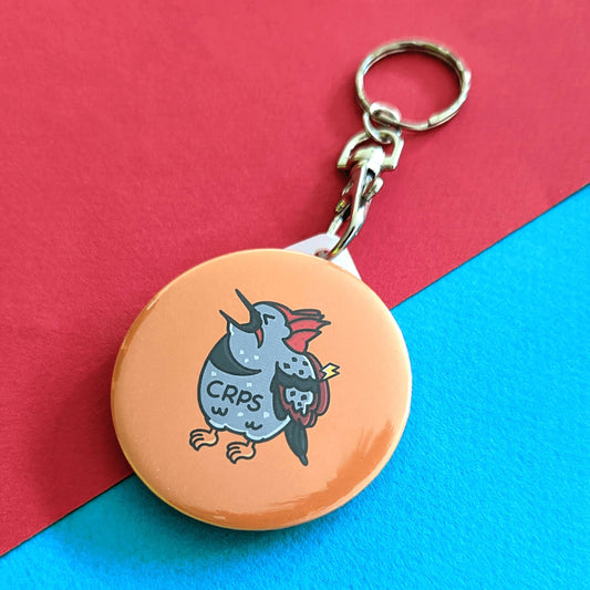 CRPS Keyring - Complex Regional Pain Syndrome on a red and blue background. The silver clip yellow plastic circular keychain features a screaming upset woodpecker bird with a lightning bolt on its wing and 'CRPS' written across its belly in black. The design is raising awareness for complex regional pain syndrome.