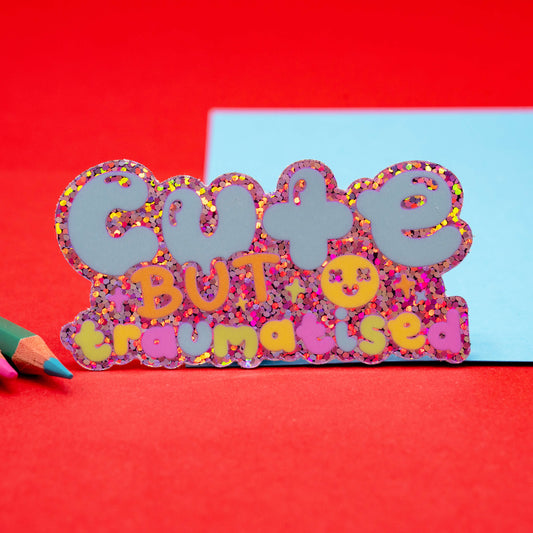 Gloss vinyl sticker by Innabox that says “Cute But Traumatised” in pastel colours on a sparkly holographic glitter background, styled on red felt and blue card with coloured pencils. Uplifting disability-themed design for neurodivergent and chronically ill communities.
