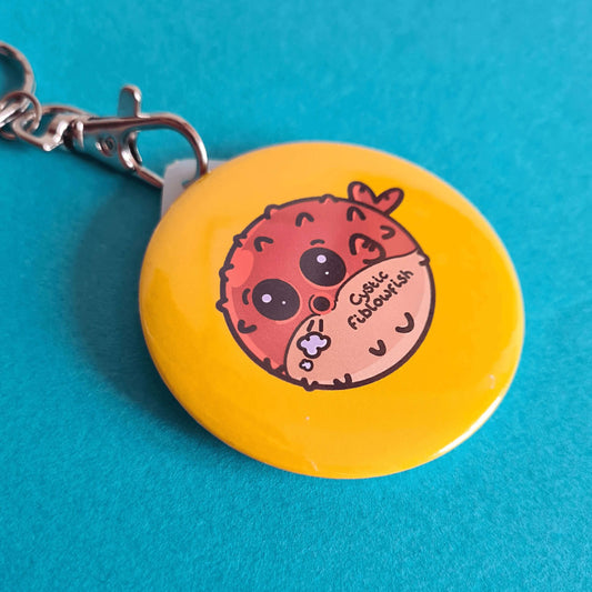 The Cystic Fiblowfish Keyring - Cystic Fibrosis on a blue background. The silver clip plastic yellow circle keychain with a puffer fish blowfish wheezing with 'cystic fiblowfish' written in black on its belly. The design is raising awareness for cystic fibrosis.