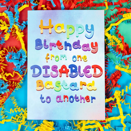 The Happy Birthday From One Disabled Bastard To Another Card on a red, blue and green background with red, yellow and blue crinkle card confetti. The pastel blue and purple a6 birthday card has rainbow bubble writing reading 'happy birthday from one disabled bastard to another'. The hand drawn design is a humorous greeting card raising awareness for disabled people. 