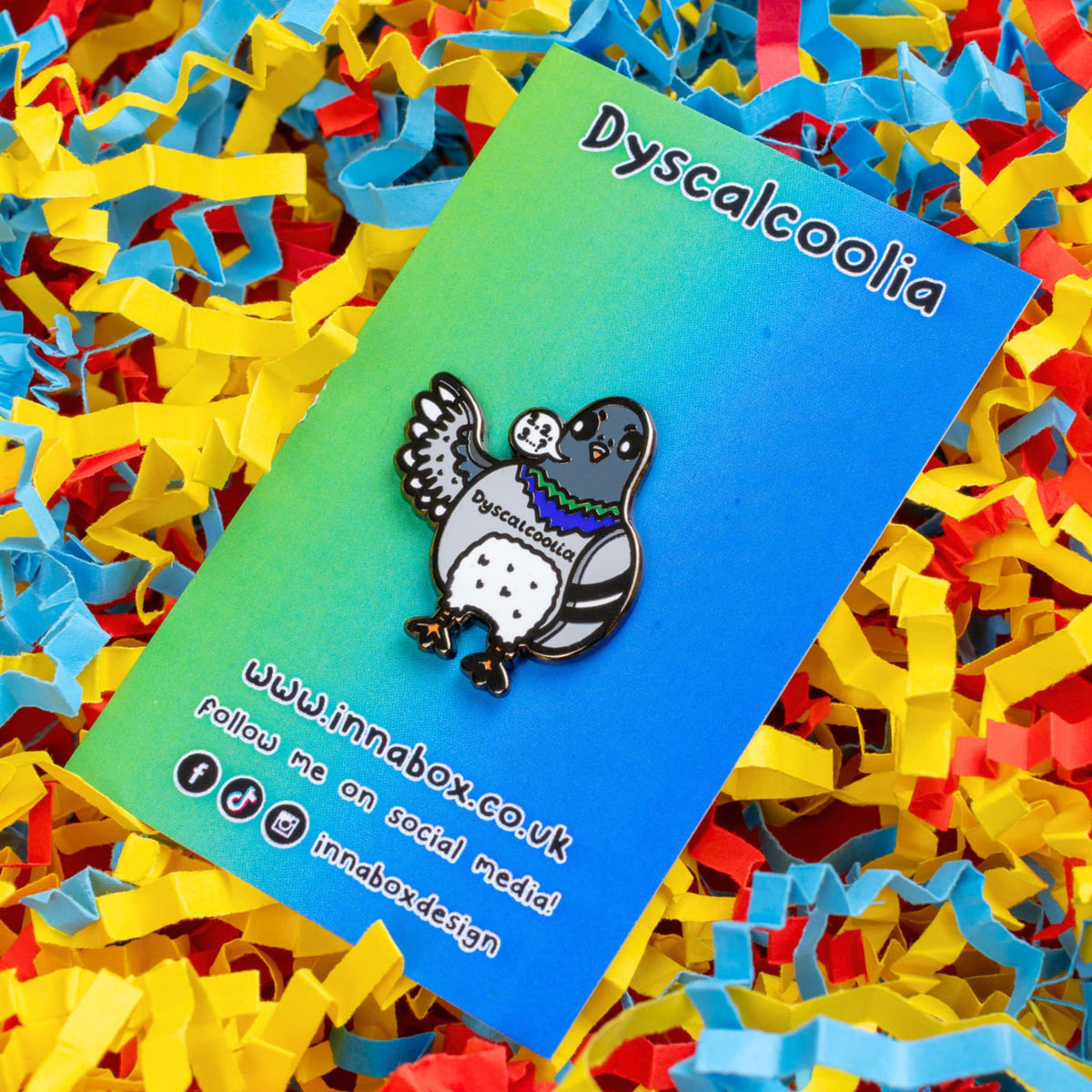 The Dyscalcoolia Pigeon Enamel Pin - Dyscalculia on a blue to green gradient backing card with black text laid on a red, yellow and blue confetti card background. A Pigeon looking confused holding up a wing with a speech bubble full of numbers and a question mark across its chest reads 'dyscalcoolia'. The enamel pin is raising awareness for dyscalculia.