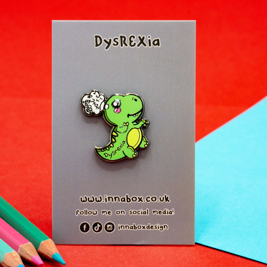 A Dysrexia Dinosaur enamel pin by Innabox displayed on a branded backing card. The green dino character has “Dysrexia” written on its side and a thought bubble with jumbled letters, symbolising dyslexia. Photographed on a bold red background with coloured pencils, promoting disability awareness through cute design.