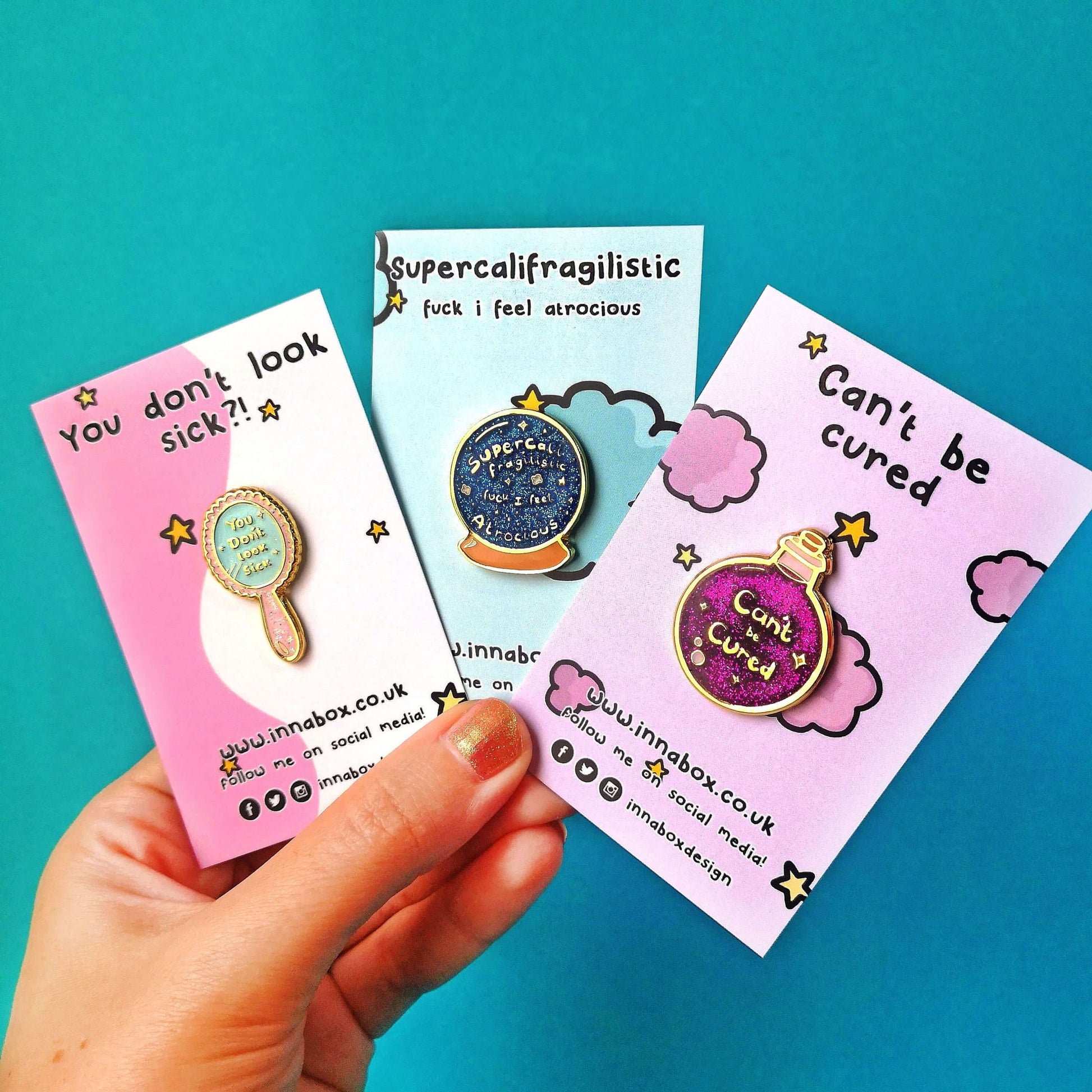Three Innabox enamel pins shown on backing card held by a hand on a blue background. Choose from any three pins. Enamel pins designed to raise awareness for chronic illnesses