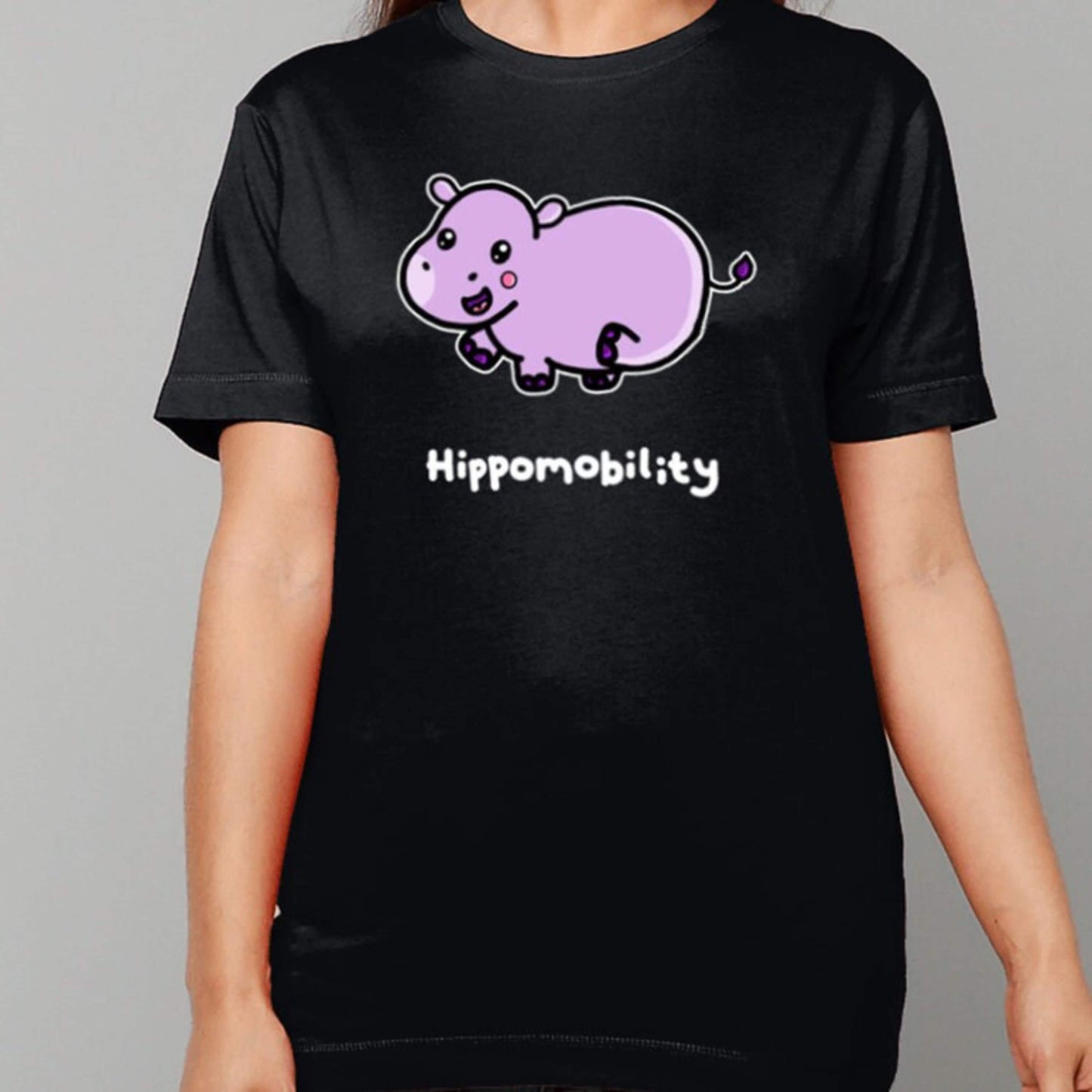 femme person wearing Hippomobility Tee - Hyper Mobility Tee standing in front of a grey backdrop. The black t-shirt has an illustration of a pink happy hippo with 'hippomobility' in white text underneath. The hand drawn design is made to raise awareness for hyper mobility.