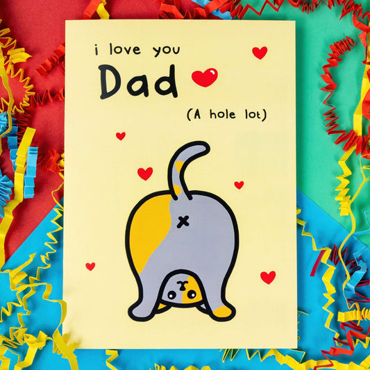 I Love You Dad - Cat Card on a red, green and blue background with coloured card confetti. The pale yellow card has black text at the top that says 'I love you Dad (a hold lot)'. There is an illustration of a grey and ginger  cat bending over to show it's bum in the air with red love hearts dotted around the card.