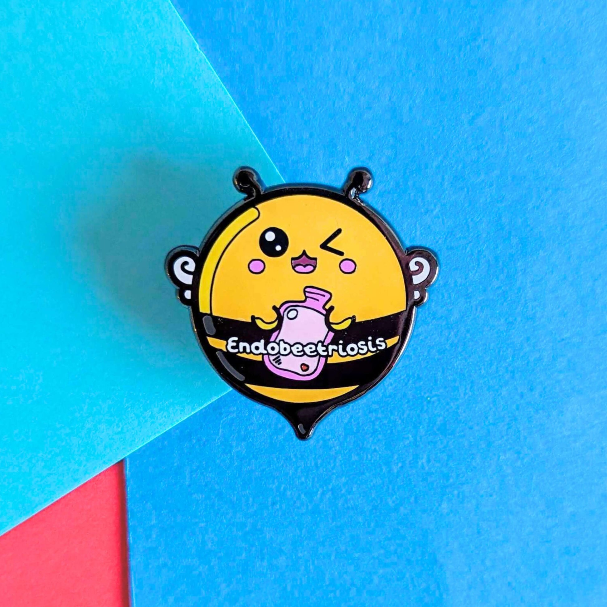 Endobeetriosis 2.0 Bee Enamel Pin - Endometriosis on a red and blue background. The enamel pin is a bumble bee with a smiley face and holding a pink hot watterbottle in its hands, across its middle is white text that reads endobeetriosis. The enamel pin is designed to raise awareness for endometriosis