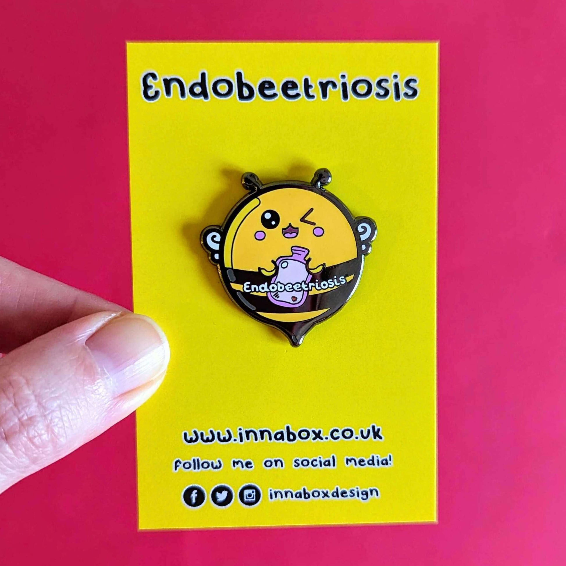 Endobeetriosis 2.0 Bee Enamel Pin - Endometriosis on yellow backing card held over a red background. The enamel pin is a bumble bee with a smiley face and holding a pink hot watterbottle in its hands, across its middle is white text that reads endobeetriosis. The enamel pin is designed to raise awareness for endometriosis