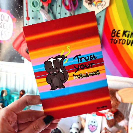 Trust Your Instinks - Skunk Postcard held in front of a decorated wall. The coloured stripy postcard has an illustration of a skunk with one eye closed and green fumes coming from it's tail. 'Trust your' is written next to it in black text with 'instinks' under it in green text