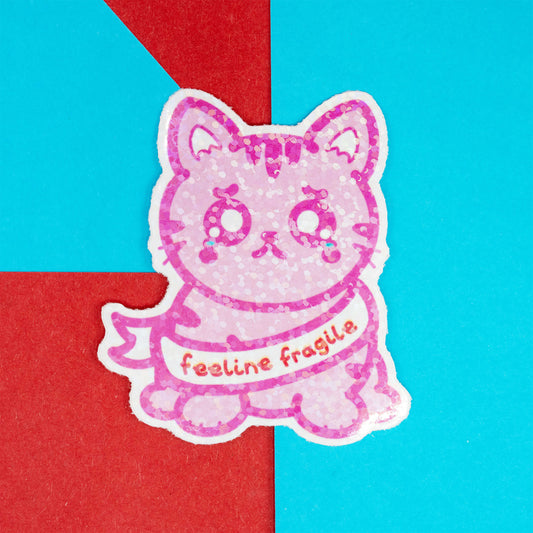 Glittery pink cat-shaped sticker with sad eyes and “feeline fragile” slogan, lying flat on a bright red and blue background. From Innabox, a UK brand raising awareness for chronic illness and hidden disabilities.