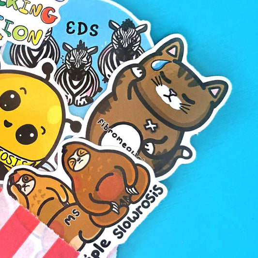 The Fibromeowgia Cat Sticker - Fibromyalgia Syndrome on a blue background amongst other innabox stickers coming out of a red stripe candy bag. The brown sad tabby cat shaped sticker has one paw on its head and the other on its tummy with a white bandaid and sweat droplet, across its middle reads 'fibromeowgia'. The hand drawn design is raising awareness for fibro fibromyalgia syndrome.