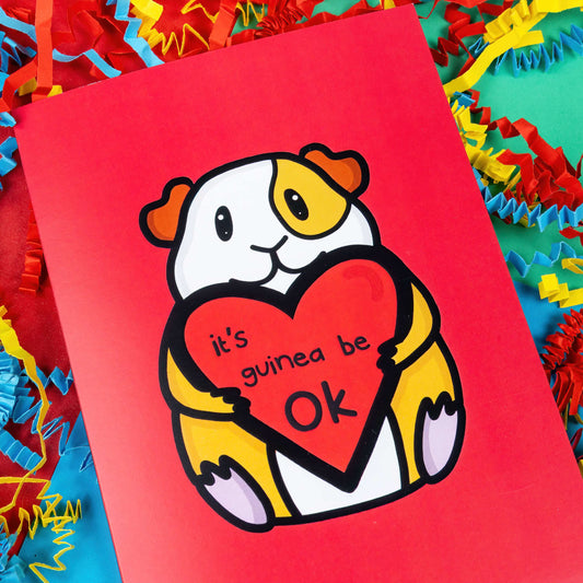 The It's Gonna Be OK Guinea Pig Card on a red, blue and green background with yellow, blue and red crinkle card confetti. The red a6 greeting card features a smiling orange and white guinea pig sat down holding a red heart with black text reading 'it's guinea be ok'. The hand drawn design is a perfect send a hug card as a gentle positive reminder.