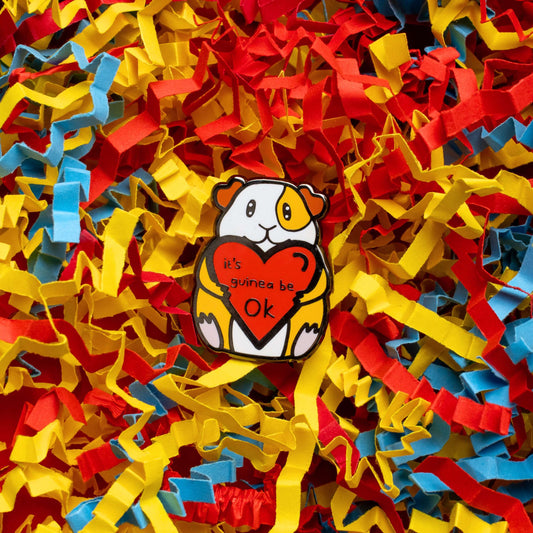 Guinea Pig Mental Health Enamel Pin shown on a red, blue and yellow card confetti background. The enamel pin is of a smiling guinea pig holding a big red heart with the text it's guinea be ok inside. The enamel pin is deigned to raise awareness for mental health.