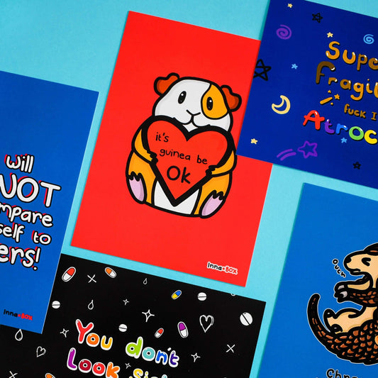 The It's Gonna Be OK Guinea Pig Postcard on a blue background with other innabox postcards. The red postcard print features a cute orange and white smiling guinea pig sat holding a red heart with black text reading 'it's guinea be ok'. The hand drawn design is a reminder for staying positive.