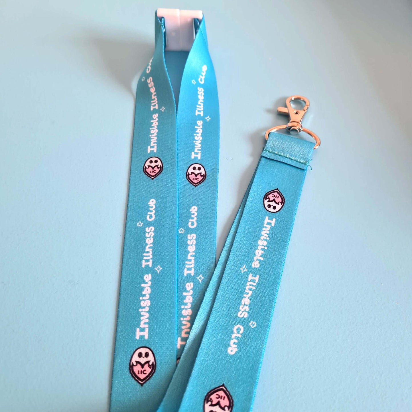 The Invisible Illness Club Lanyard on a pastel blue background. The pastel blue lanyard has repeating white text reading 'invisible illness club' and innabox pastel ghost logo. It has a silver lobster clip and white safety break. The hand drawn design is raising awareness for hidden disabilities.