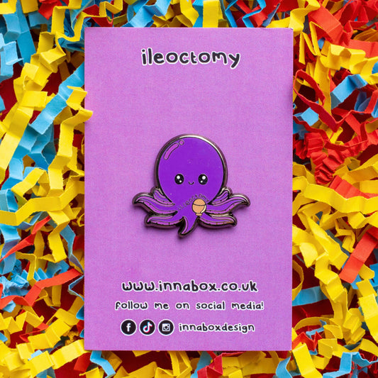 Ileoctomy Enamel Pin - Ileostomy on purple backing card laid on a yellow, blue and red card confetti background. The enamel pin is a cute smiling purple octopus sticker with text saying ileoctomy on its belly with a stoma bag underneath. Enamel pin designed to raise awareness for Ileostomy.