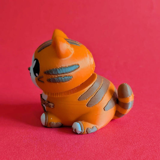 Side profile of Innabox Neurodiverse Kitty Desk Buddy showing striped tail, layered 3D printed texture and rounded shape. Orange and brown sensory fidget toy with clicker head movement, palm sized at approx 8 x 7 x 6 cm. Biodegradable polylactic acid desk buddy for autism, anxiety and neurodivergent support.