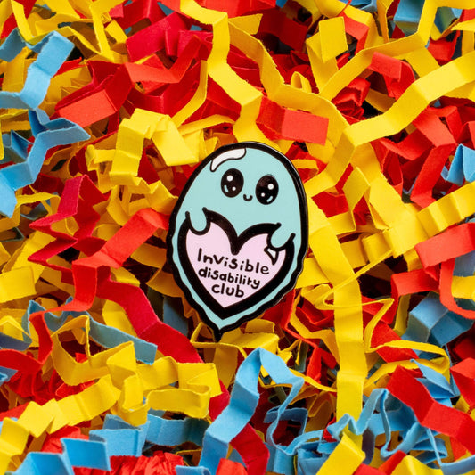 Invisible Disability Club Ghost Enamel Pin shown on a red, blue and yellow confetti card background. The enamel pin is of a cute smiling ghost holding a pink heart with text saying invisible disability club. The enamel pin is designed to raise awareness for hidden and chronic disabilities.
