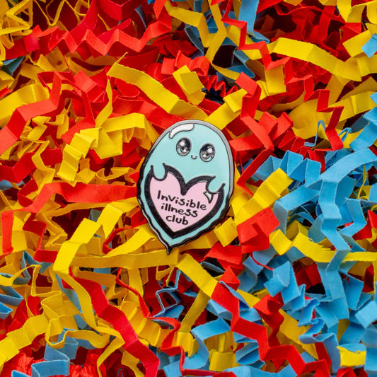 Invisible Illness Club Enamel Pin on a red, blue and yellow crinkle card confetti background. The enamel pin is of a cute smiling ghost holding a pink heart with text saying invisible illness club. The enamel pin is designed to raise awareness for hidden and chronic illnesses.