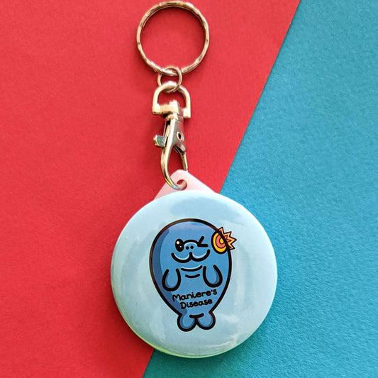 The Maniere's Disease Manatee Keyring - Ménière's Disease on a red and blue background. The pastel blue circular plastic keychain with silver lobster clip features a winking manatee with a yellow and red inflamed ear and black text across its body reading 'maniere's disease'. The hand drawn design is raising awareness for Ménière's Disease and vertigo.