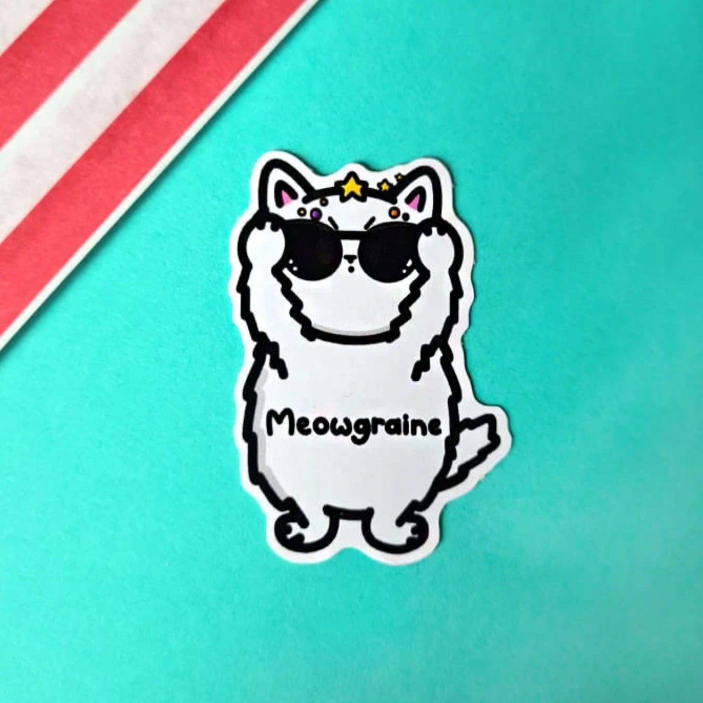 The Meowgraine Cat Sticker - Migraine on a blue background with a red stripe candy bag. A white stressed cat sticker clutching a pair of black sunglasses to its eyes with multicoloured spots and stars over its head, across its middle reads 'meowgraine'. The hand drawn design is raising awareness for migraines and headaches.
