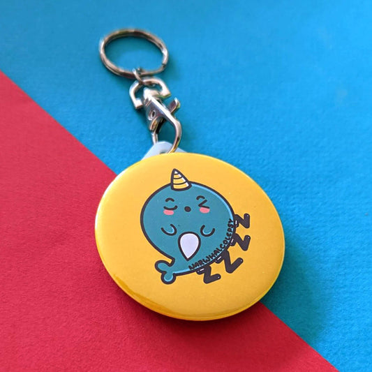 The Narwhalcolepsy Narwhal Keyring - Narcolepsy on a red and blue background. A silver lobster clip with a yellow plastic circular keychain of a sleeping blue narwhal on a bed of ZZZ with black text across its body reading 'narwhalcolepsy'. The hand drawn design is raising awareness for narcolepsy.