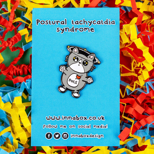 An enamel pin of a cat illustration with spinning dizzy stars around its head with its paws outstretched and postural tachycatdid syndrome (pots) written on its belly on blue backing card on a coloured card confetti background. Postural tachycardia syndrome (PoTS) is when your heart rate increases very quickly after getting up from sitting or lying down.