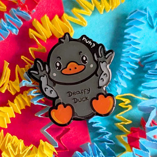 Close-up of Deaffy Duck enamel pin by Innabox against a colourful textured background. The grey duck character signs BSL with both hands raised, featuring orange beak and feet and a playful “huh?” detail. A cheerful enamel pin celebrating Deaf culture and disability awareness.