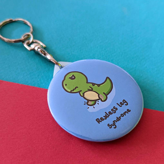 The Rexless Leg Syndrome Keyring - Restless Leg Syndrome on a red and blue background. The silver lobster clip plastic blue circular keychain features an angry upset green t-rex dinosaur with red lower legs and black lines, underneath in black text reads 'rexless leg syndrome'. The hand drawn design is raising awareness for restless legs syndrome.