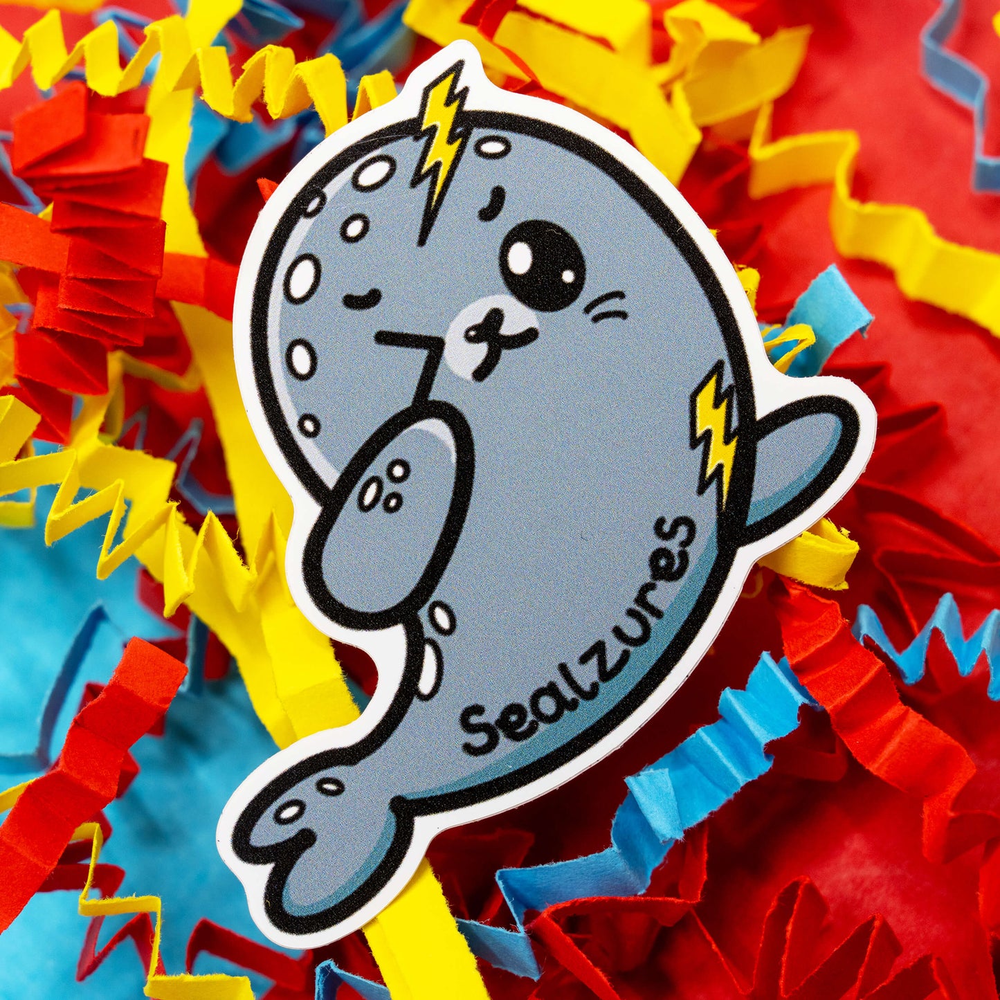 Adorable Sealzures sticker by Innabox featuring a cute grey seal with lightning bolts and a playful expression to represent seizure awareness. Surrounded by colourful confetti, this waterproof gloss vinyl sticker is approx 5 cm tall and custom cut. A kind and funny design to support those living with epilepsy or hidden disabilities.