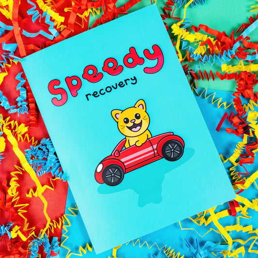 The Speedy Recovery Get Well Soon Cat Card on a colourful card confetti background. The blue card has 'speedy' written at the top in big red letters with 'recovery' written in a smaller black font underneath. In the centre of the card is an adorable orange cat with an open mouthed smile to reveal it's pink tongue. The cat has pink ears and nose and big eyes. The cat is driving a red car.