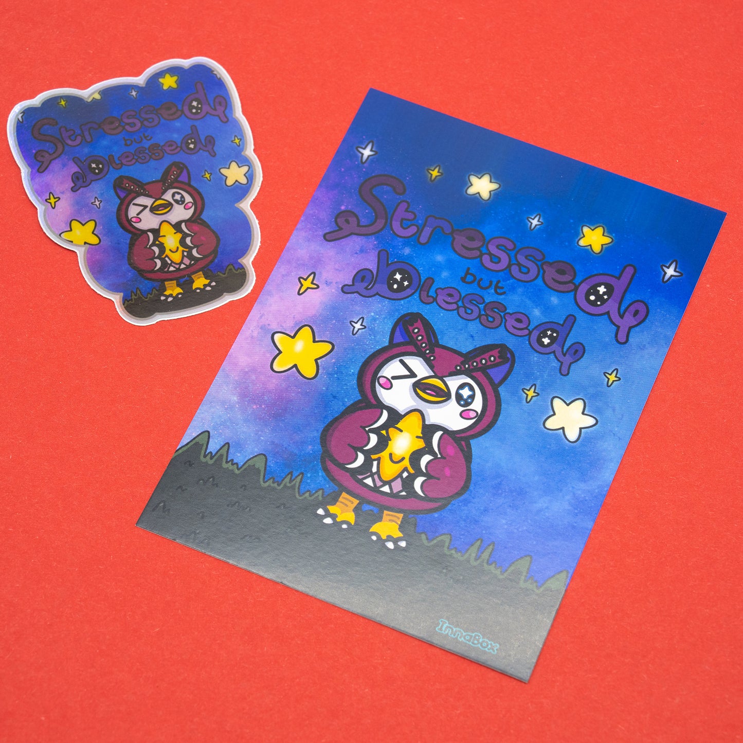 A holographic Stressed But Blessed vinyl sticker by Innabox placed beside a matching art print. The cute owl-style character holds a glowing star on a galaxy background with yellow stars and a custom cut border. Displayed on a red background to show the vibrant colours and holographic shine. A comforting design for disabled and chronically ill folk.