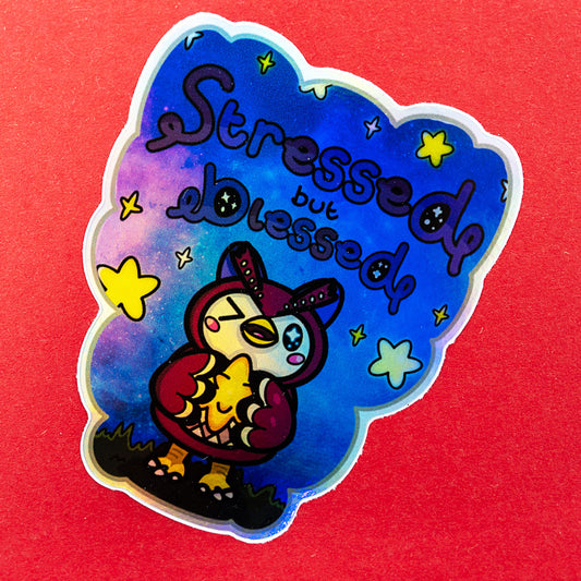 A holographic Innabox vinyl sticker showing a sweet owl-like character hugging a bright star under the quote Stressed But Blessed. The sticker has a colourful galaxy background, yellow stars and a custom cut outline. Photographed on a red surface to highlight the holographic shine. Ideal for uplifting chronic illness and disability communities.