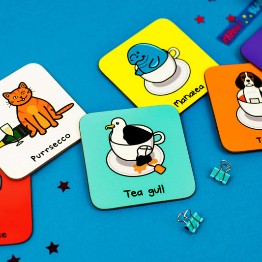 Tea Gull Seagull Coaster on a blue background with other innabox coasters and sequins. The aqua blue wooden coaster has an illustration of a seagull inside of a tea cup and saucer with a used tea bag on the side.