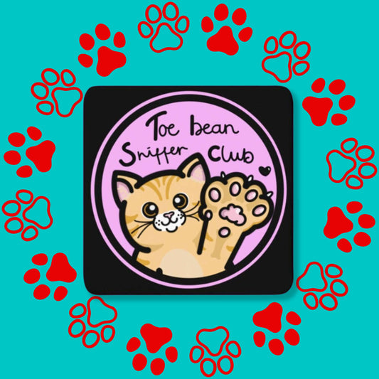 The Toe Bean Sniffer Club Coaster on a red and blue paw print background. The black wooden coaster with rounded edges has a pastel pink circle with black text reading 'toe bean sniffer club' with an orange smiling cat lifting its paw up with a black heart.