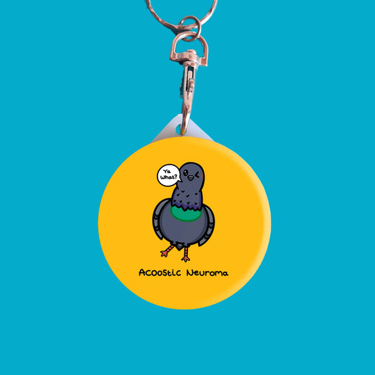A yellow circular keyring with a grey pigeon with a green chest on the front. The pigeon has one eye closed and one leg sticking out to the side. There is a speechbubble coming from it's beak that reads 'ya what?''. 'Acoostic Neuroma' is written underneath. The background of the photo is blue. Raising awareness for acoustic neuroma.
