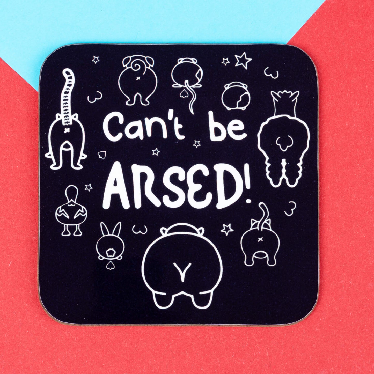 A square black coaster reading “Can’t be ARSED!” with adorable illustrated animal bums, designed by Innabox to bring humour and joy to those with invisible illnesses. Set on a bold red and blue flat-lay background.