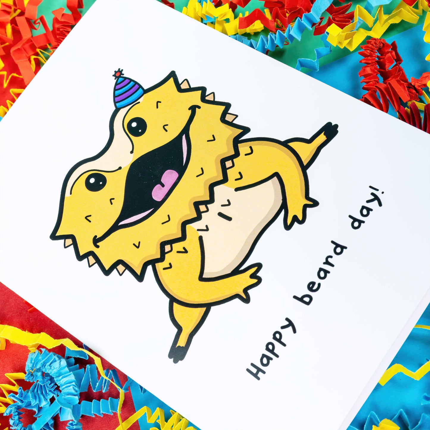 Close up of the Happy Beard Day - Bearded Dragon A6 Birthday Card on a green, blue and red background with yellow, blue and red crinkled card confetti. The white card features a happy bearded dragon reptile wearing a purple stripe party hat, underneath in black reads Happy Beard Day!