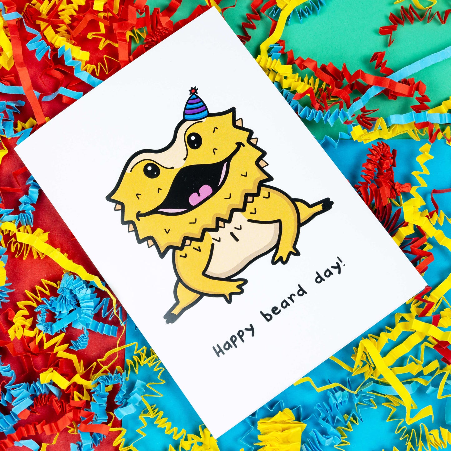 Happy Beard Day - Bearded Dragon A6 Birthday Card on a green, blue and red background with yellow, blue and red crinkled card confetti. The white card features a happy bearded dragon reptile wearing a purple stripe party hat, underneath in black reads Happy Beard Day!